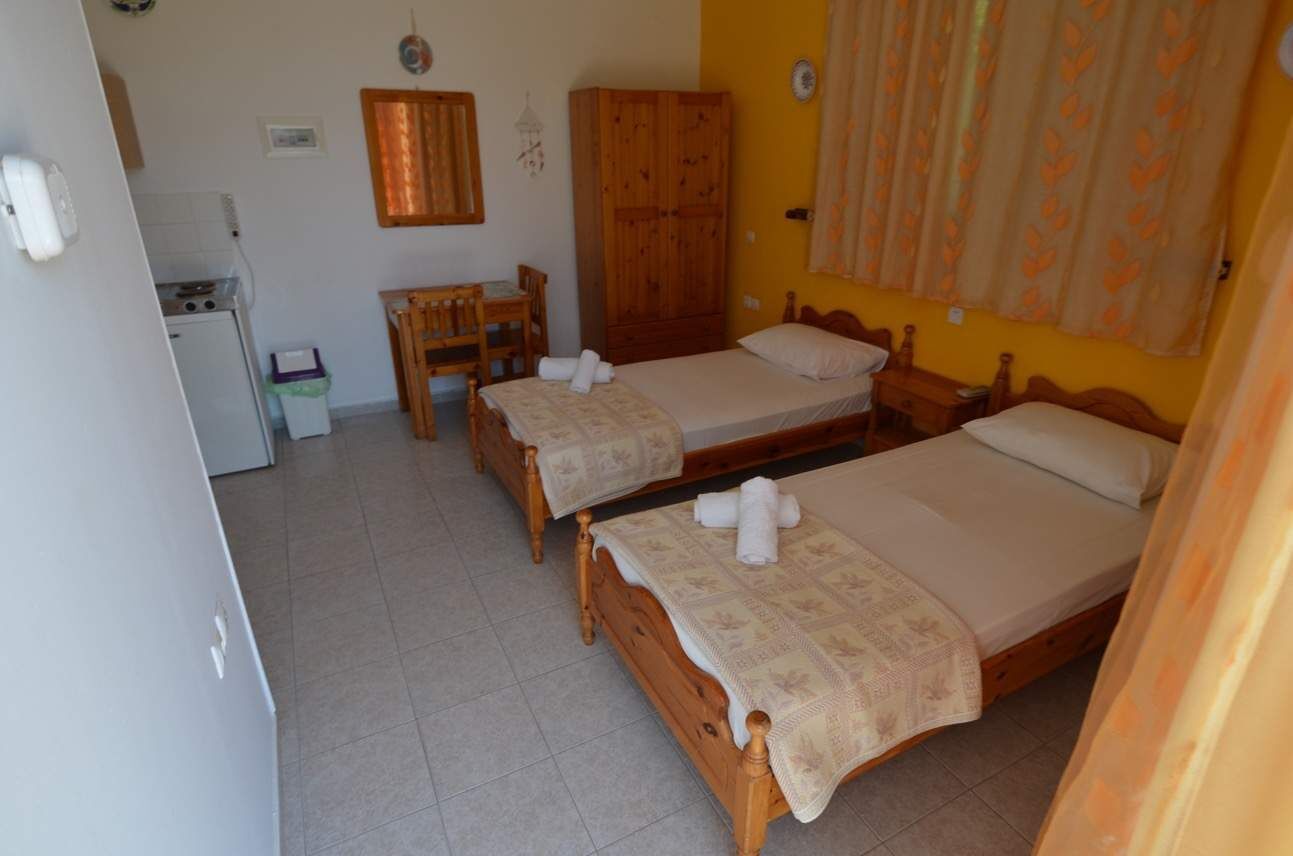 A room with two wooden beds, a wooden wardrobe, a small wooden table, a chair, a refrigerator, and a window with curtains.