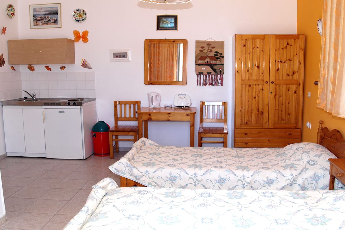 Un dormitorio con cama, armario de madera, escritorio con sillas, una pequeña cocina y baño.