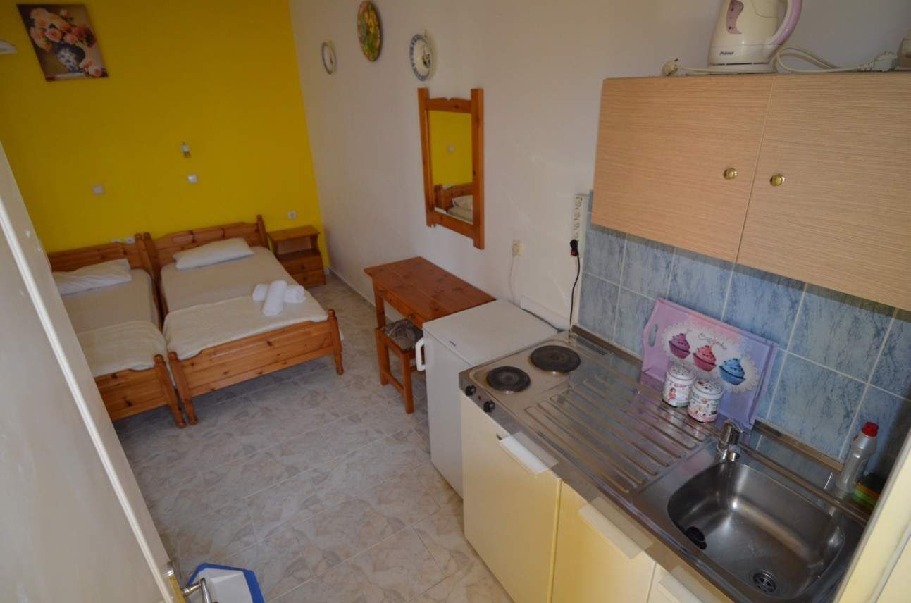 A compact room with two wooden beds, a small table, a kitchenette with a stove and sink, and a mirror on the wall.