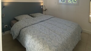 Ein Schlafzimmer mit Bett, Nachttisch, Wandlampe, Fenster mit Blick auf Grünflächen und Deckenleuchte.