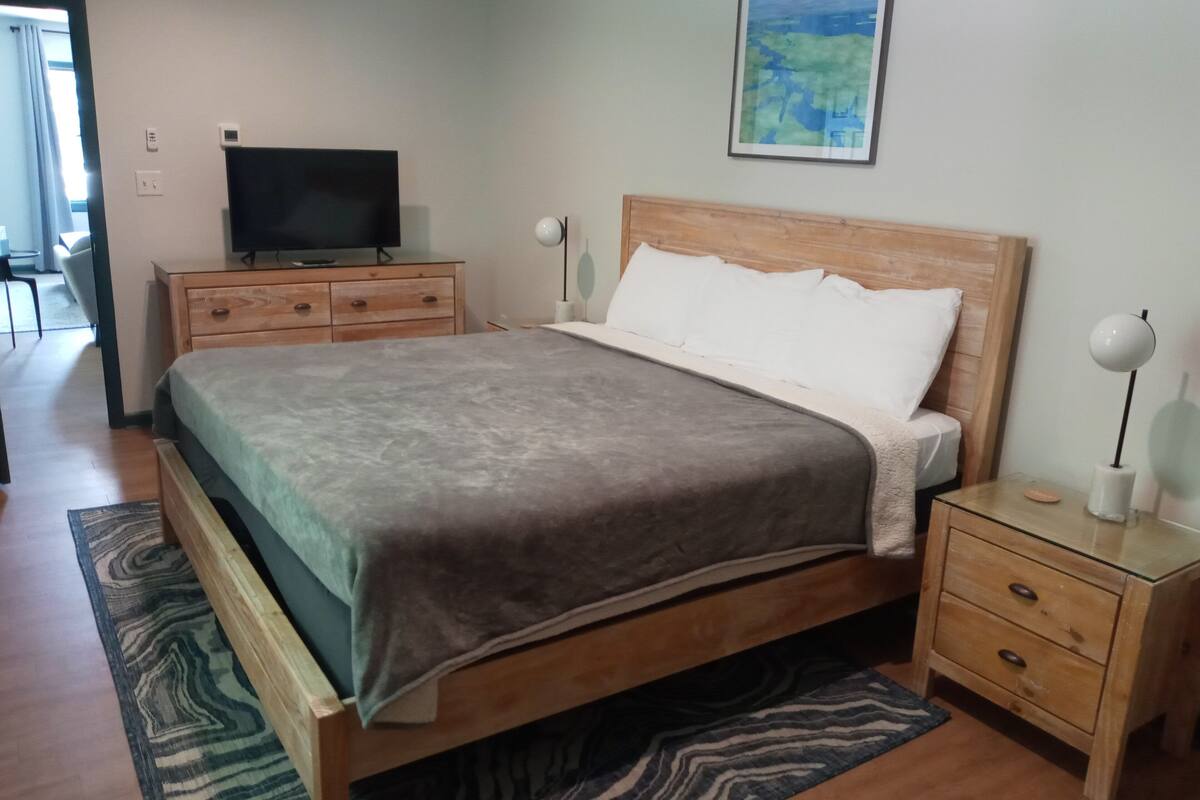 A bedroom with a wooden bed, a television on a wooden dresser, a nightstand with a lamp, and a painting on the wall.