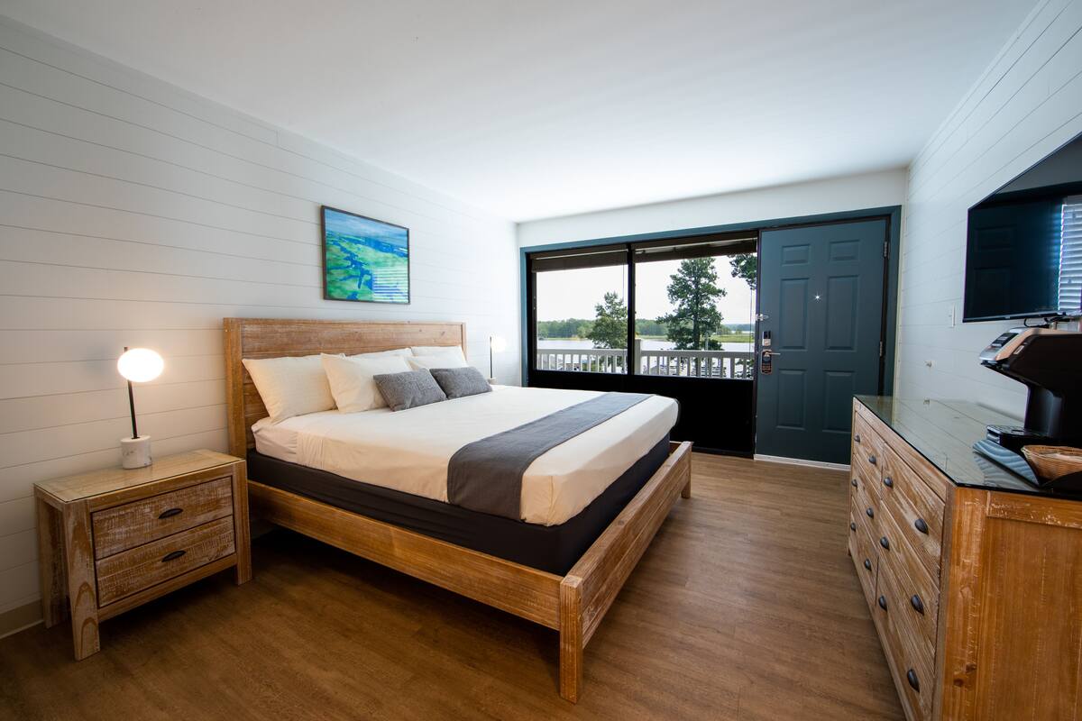 A bedroom with a wooden bed, a nightstand with a lamp, a dresser, and a large window with a view of the outdoors.