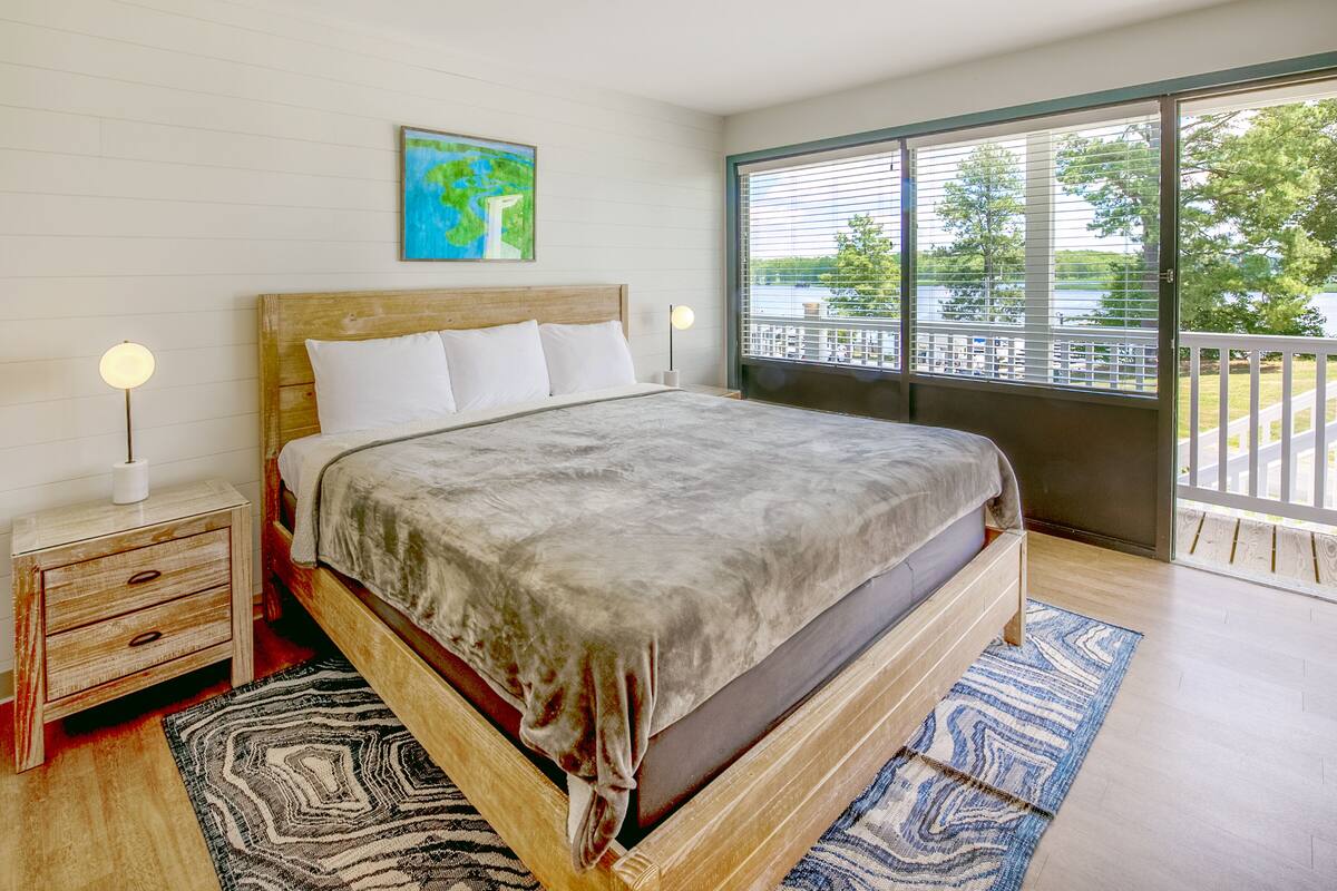 A bedroom with a large bed, a nightstand with a lamp, a painting on the wall, and a view of a balcony with a railing.