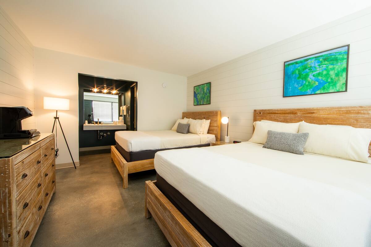 A hotel room with two single beds, a wooden dresser, a desk with a lamp, and a large abstract painting on the wall.