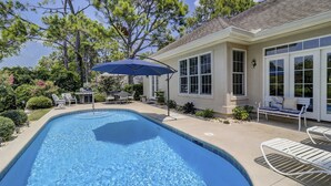 Una piscina con sillones de descanso y una sombrilla, rodeada de una casa con grandes ventanales y un patio.