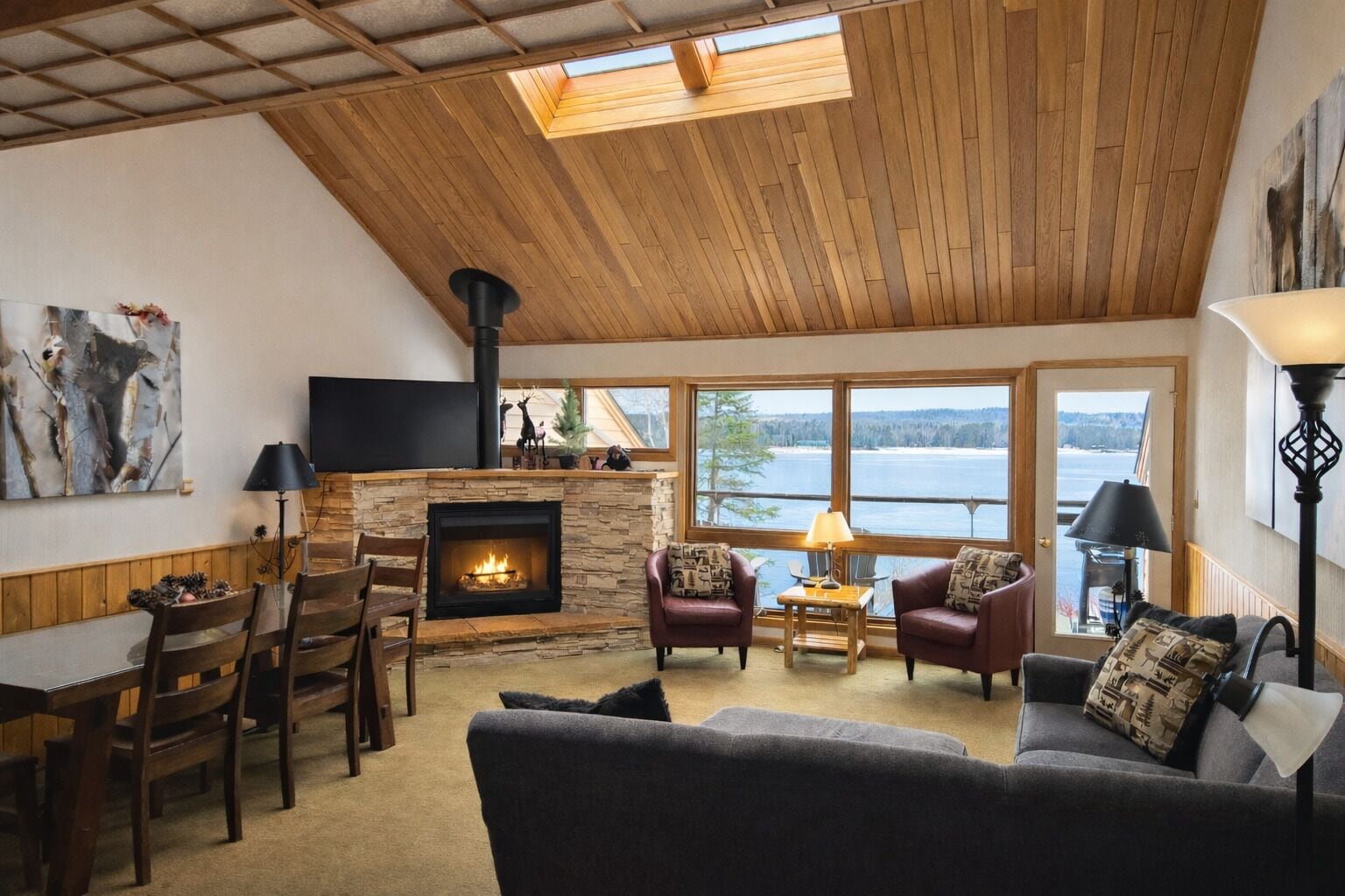 A cozy living room with a fireplace, wooden ceiling, and large windows overlooking a body of water.