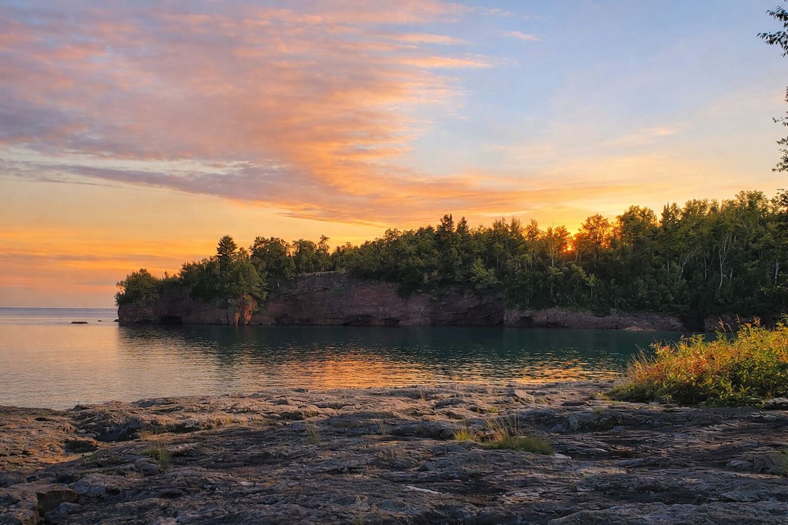 A coastal landscape with a rocky shoreline, calm water, and a forested cliff.