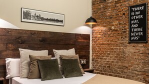 Ein Schlafzimmer mit Backsteinwand, einem Bett mit vielen Kissen, einem gerahmten Bild an der Wand und einem Schriftzug.