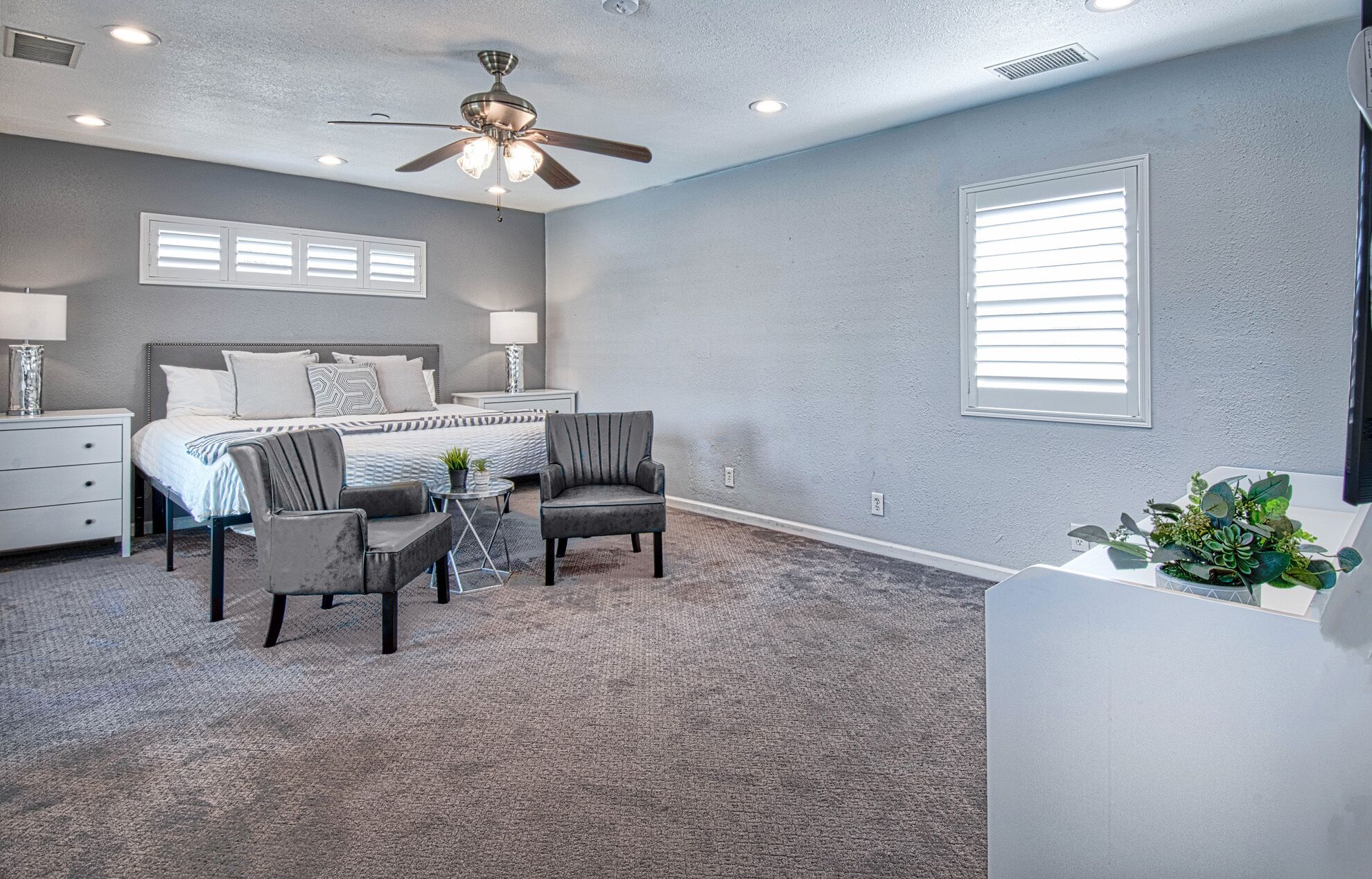 A bedroom with a bed, two armchairs, a small table, a ceiling fan, and a window with blinds.