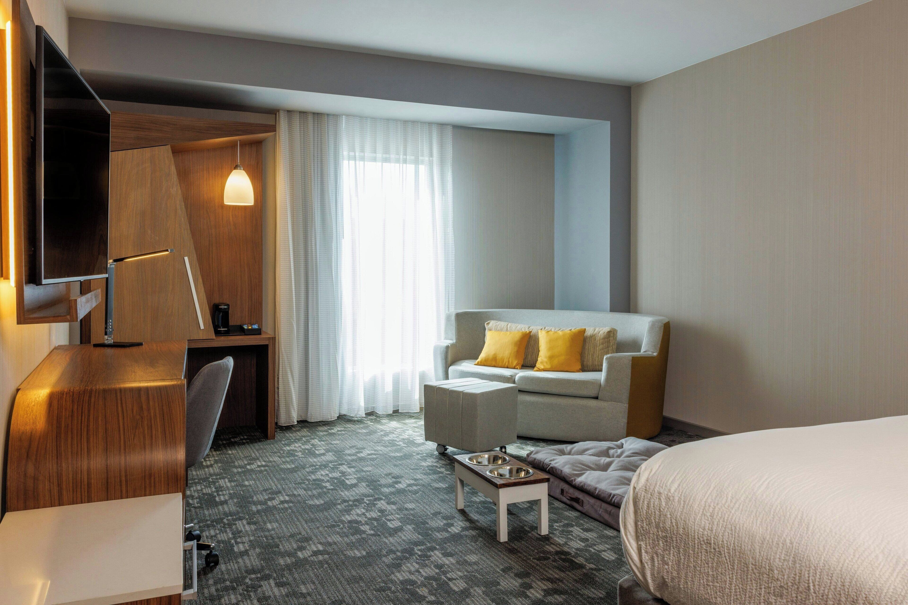 A hotel room with a bed, a sofa, a desk, a chair, a coffee table, and a TV mounted on the wall.