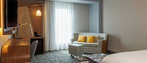 A hotel room with a bed, a sofa, a desk, a chair, a coffee table, and a TV mounted on the wall.