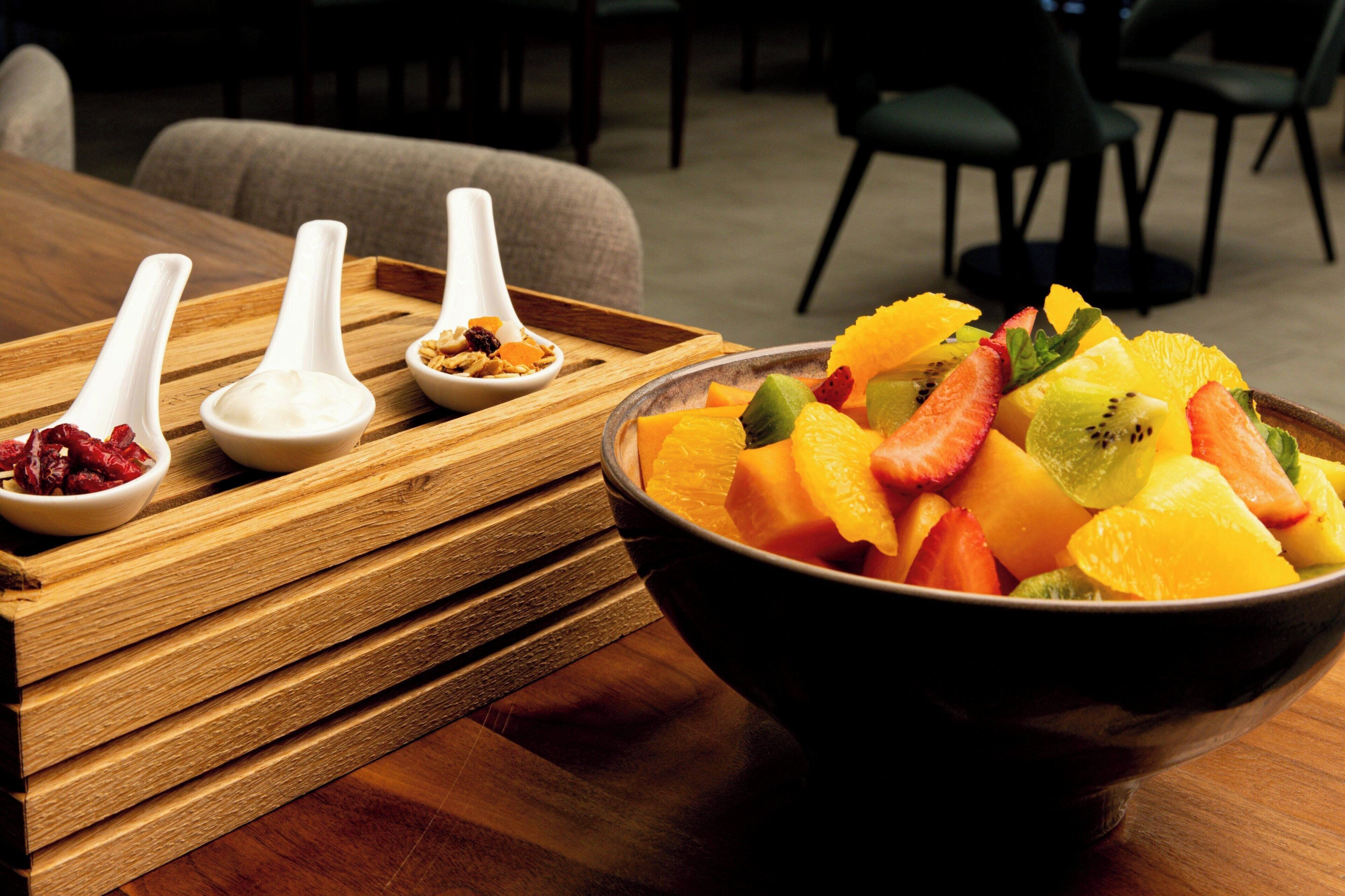 Una mesa de madera con un bol de fruta variada y tres cucharas blancas con diferentes alimentos.