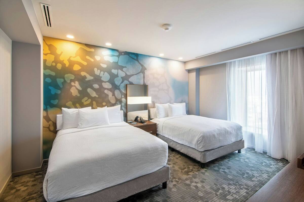 A hotel room with two beds, a large mirror, a nightstand, and a window with sheer curtains.
