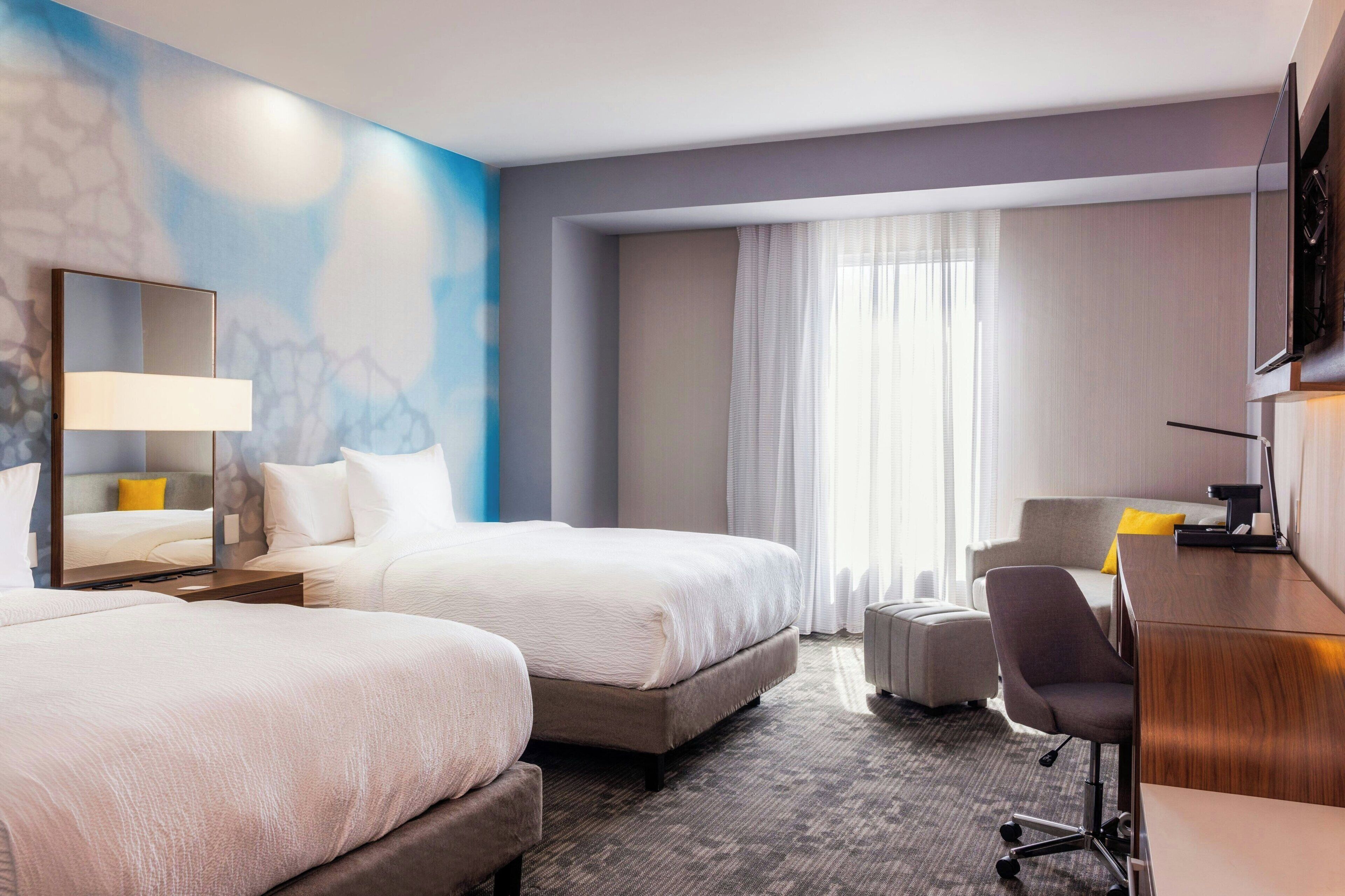 Habitación de hotel con dos camas, un escritorio, una silla, un espejo grande y una ventana con cortinas.