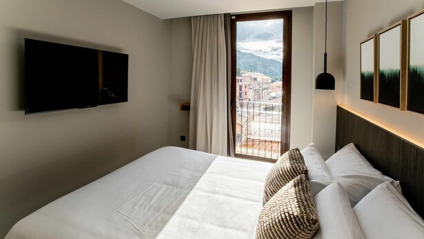 A hotel room with a large bed, a flat-screen TV mounted on the wall, a window with curtains, and a view of buildings and mountains. - Mocho- Rural- Home Villanueva A (Beceite)