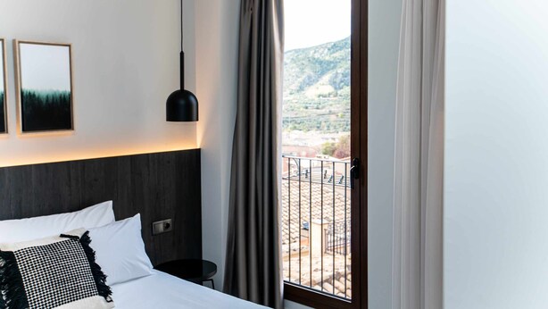 A hotel room with a bed, a bedside table, a hanging lamp, and a window with curtains. - Mocho-rural-home Lonja B (Beceite)