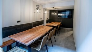 A modern dining area with wooden tables and chairs, pendant lighting, and a flat-screen TV mounted on the wall.