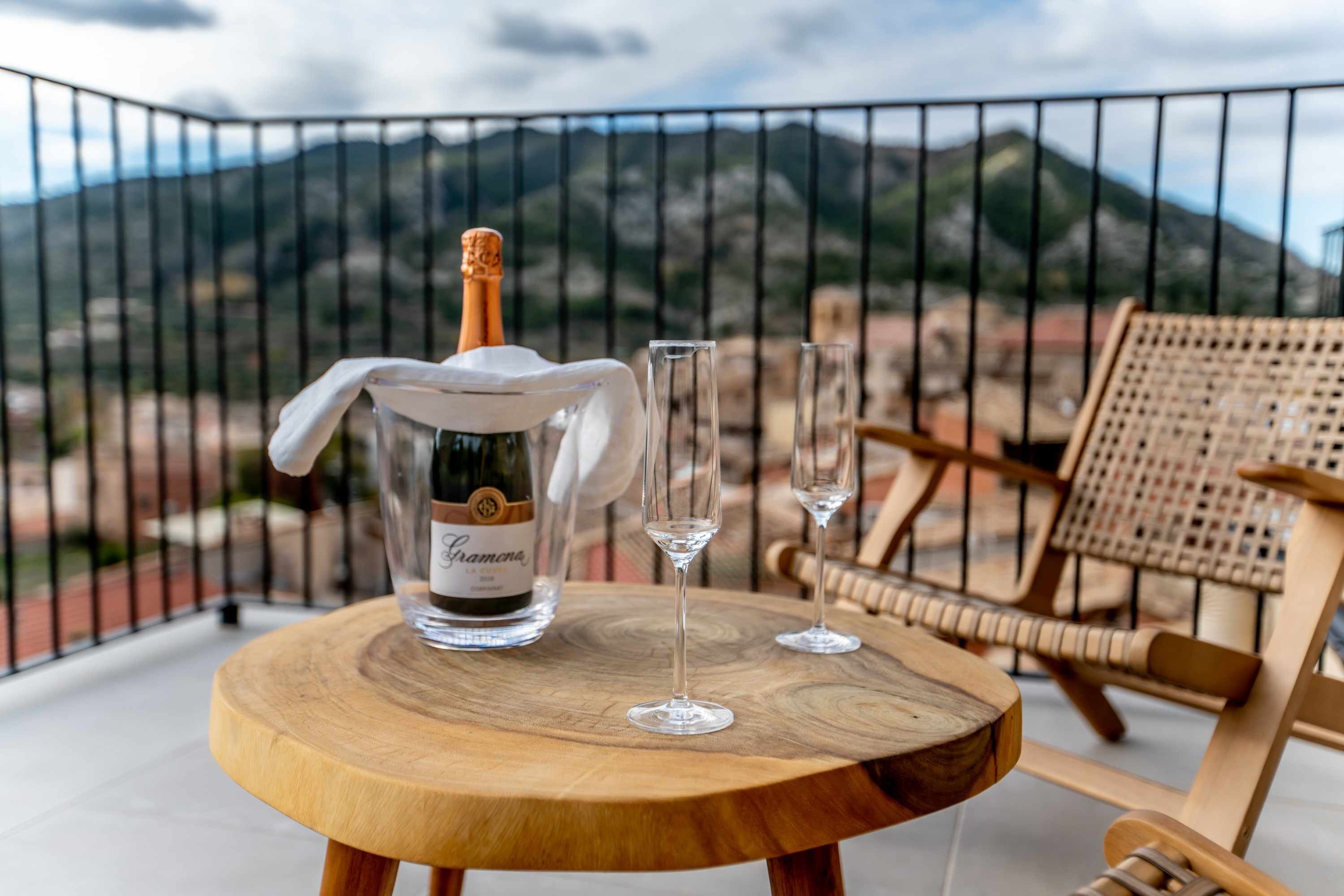 Une table en bois, une bouteille de champagne, deux verres et une chaise sur un balcon offrant une vue sur la montagne.