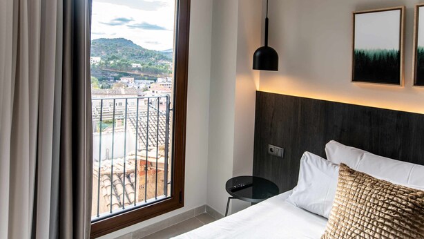 A hotel room with a large window, a bed with white linens, a round black side table, and two framed pictures on the wall. - Mocho-rural-home Lonja A (Beceite)