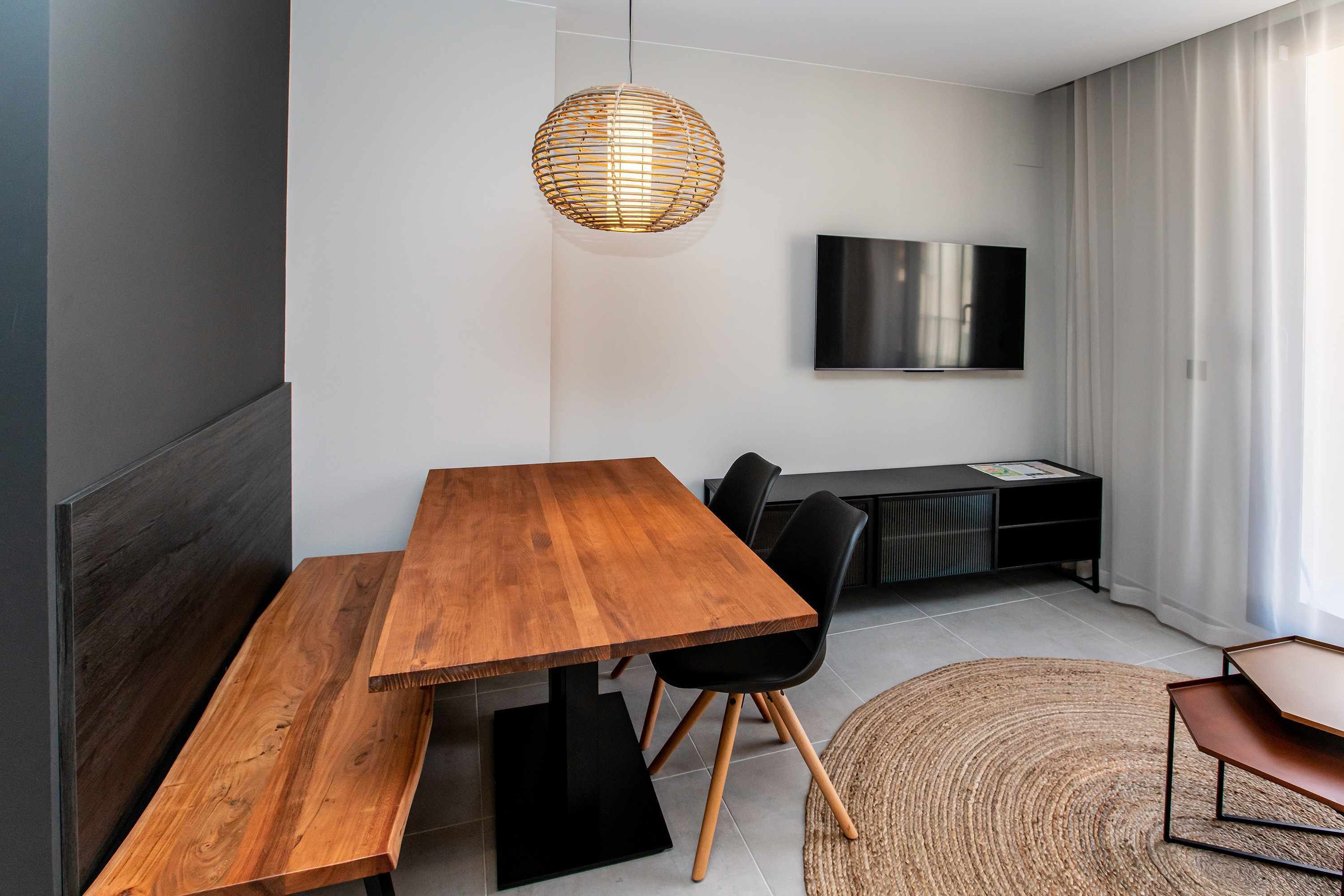 A modern dining area with a wooden table and benches, a woven pendant light, a flat-screen TV mounted on the wall, and a black entertainment unit.