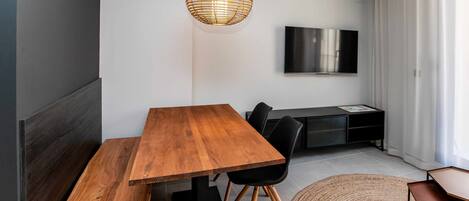 A modern dining area with a wooden table and benches, a woven pendant light, a flat-screen TV mounted on the wall, and a black entertainment unit.