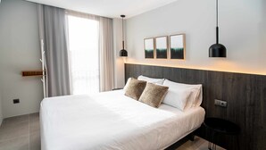 A modern hotel room with a large bed, a wooden headboard, a small side table, and a view of the outdoors through a window with curtains. - Mocho-rural-home Lonja A (Beceite)
