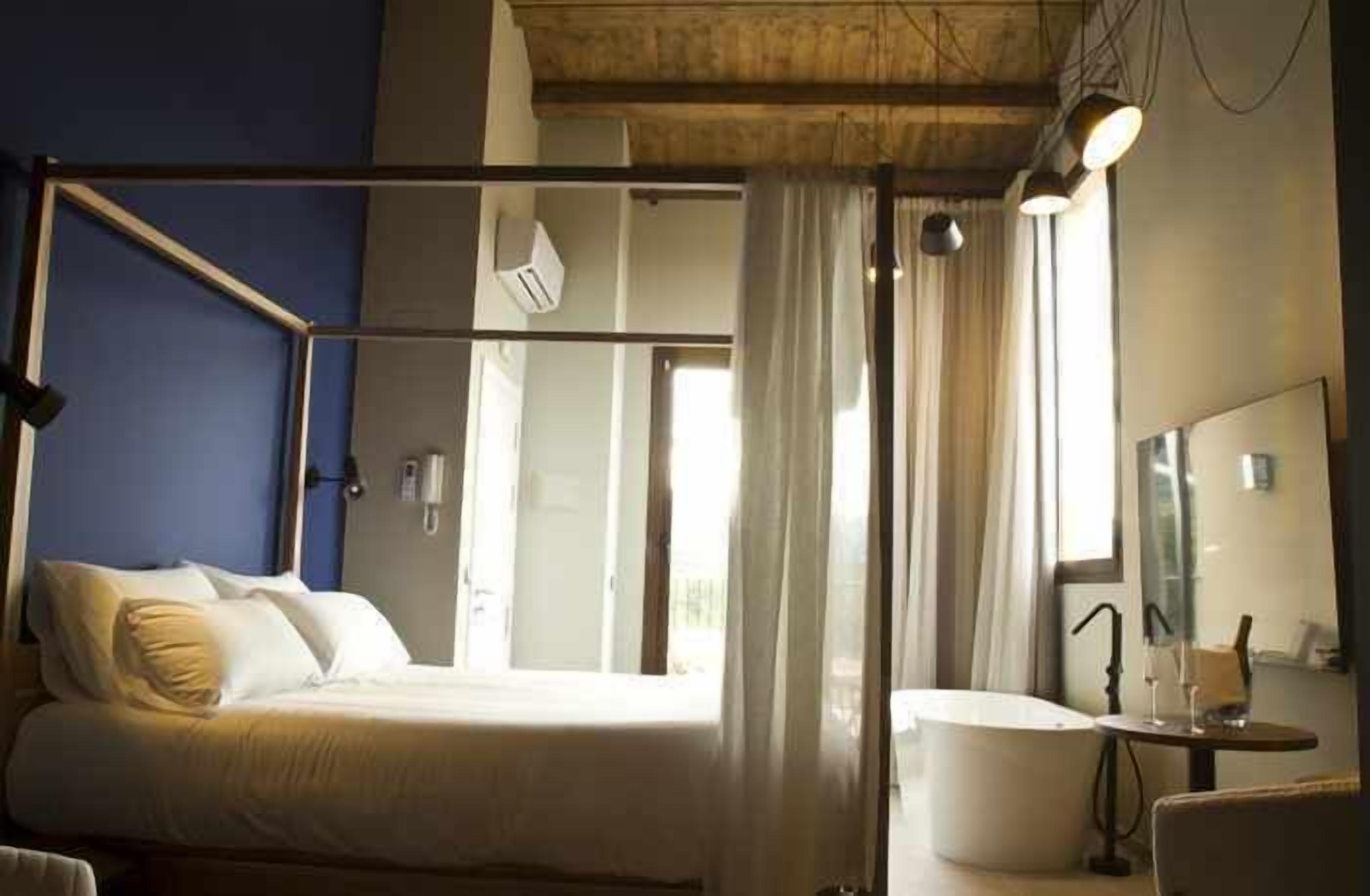 Un dormitorio con cama, mesita de noche, espejo y ventana con cortinas.