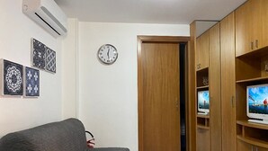 Olohuoneessa on harmaa sohva, puinen kaappi, jossa on televisio, ja seinällä oleva kello.
