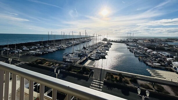 Una marina con imbarcazioni ormeggiate lungo il lungomare, un ponte sullo sfondo e cielo sereno.