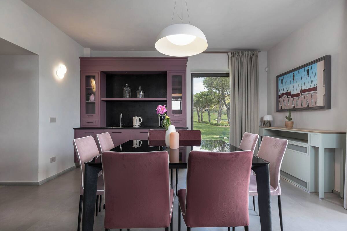 Un comedor con una mesa de vidrio, sillas rosadas, un armario morado y un cuadro en la pared.