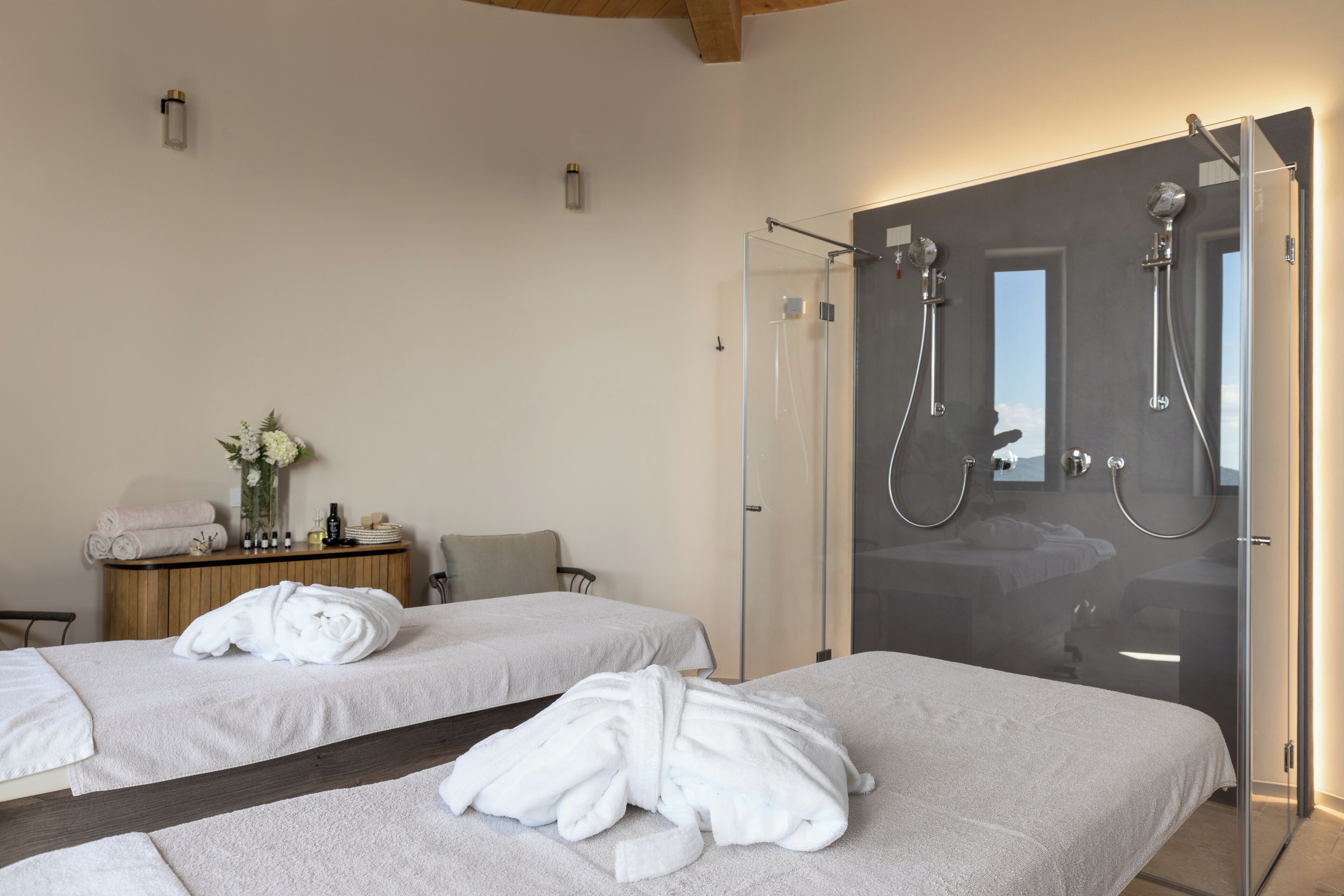 A hotel room with two beds, a glass shower enclosure, a vanity with toiletries, and a small desk with a vase of flowers.
