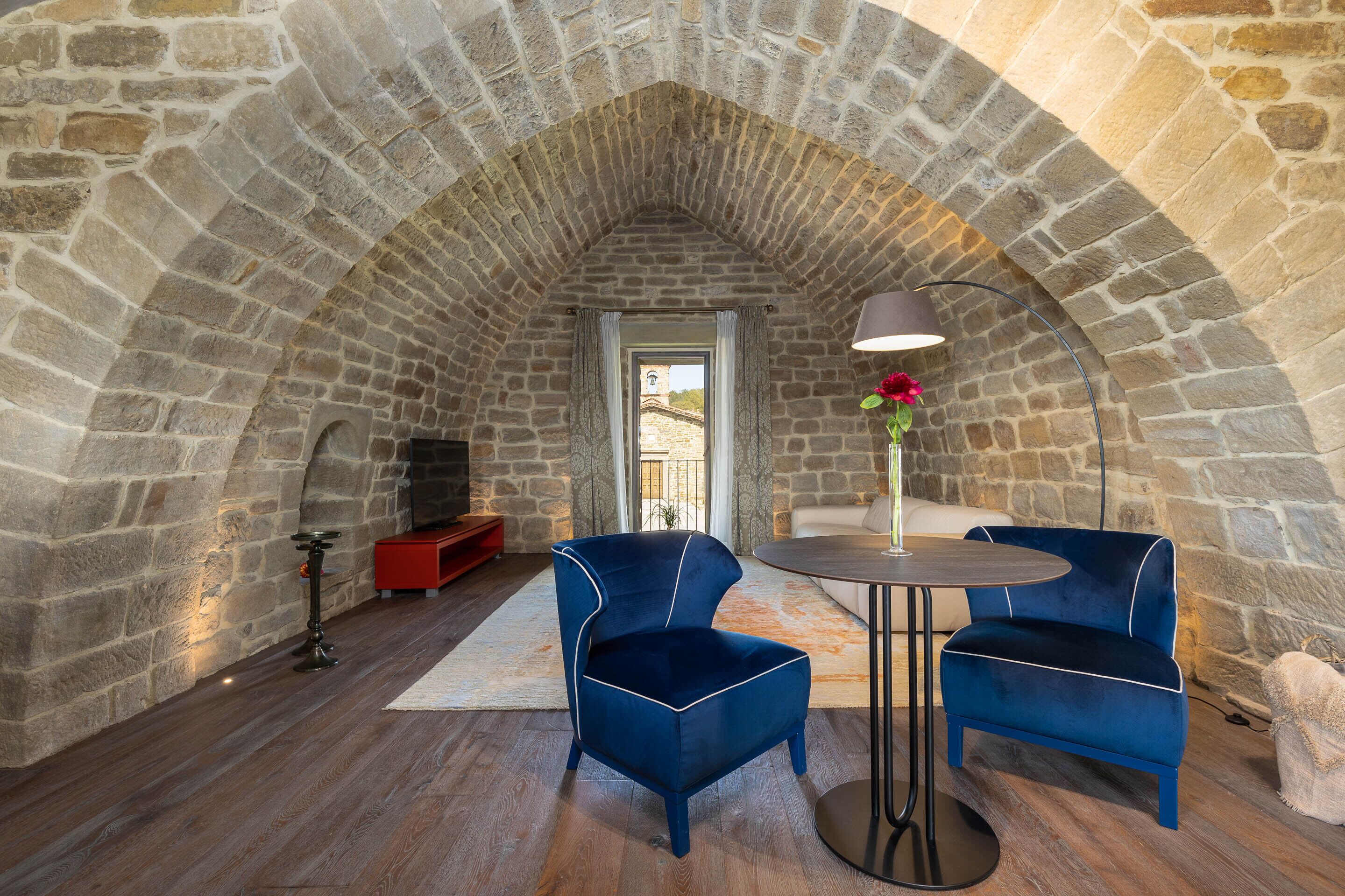A stone-walled room with a vaulted ceiling, two blue upholstered chairs, a round table, and a red cabinet.