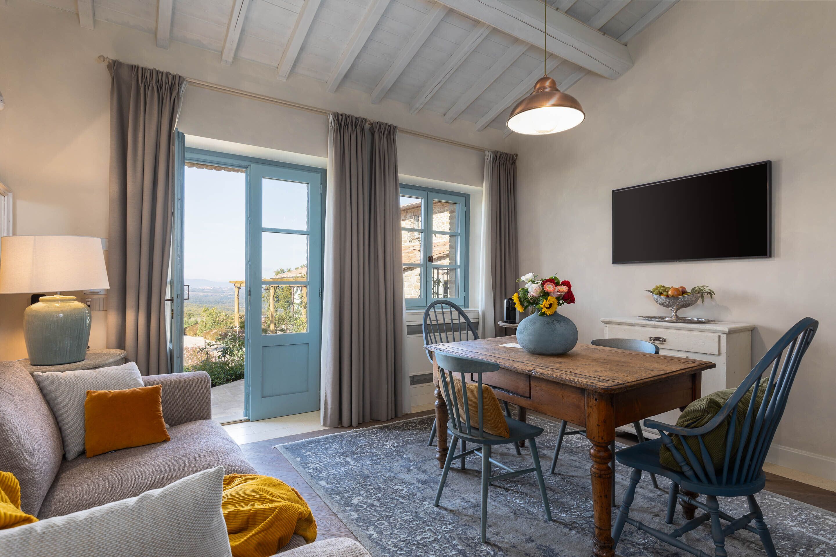 Uma sala de estar acolhedora com sofá, mesa de jantar de madeira e cadeiras. Há uma televisão fixada na parede e uma janela com cortinas.