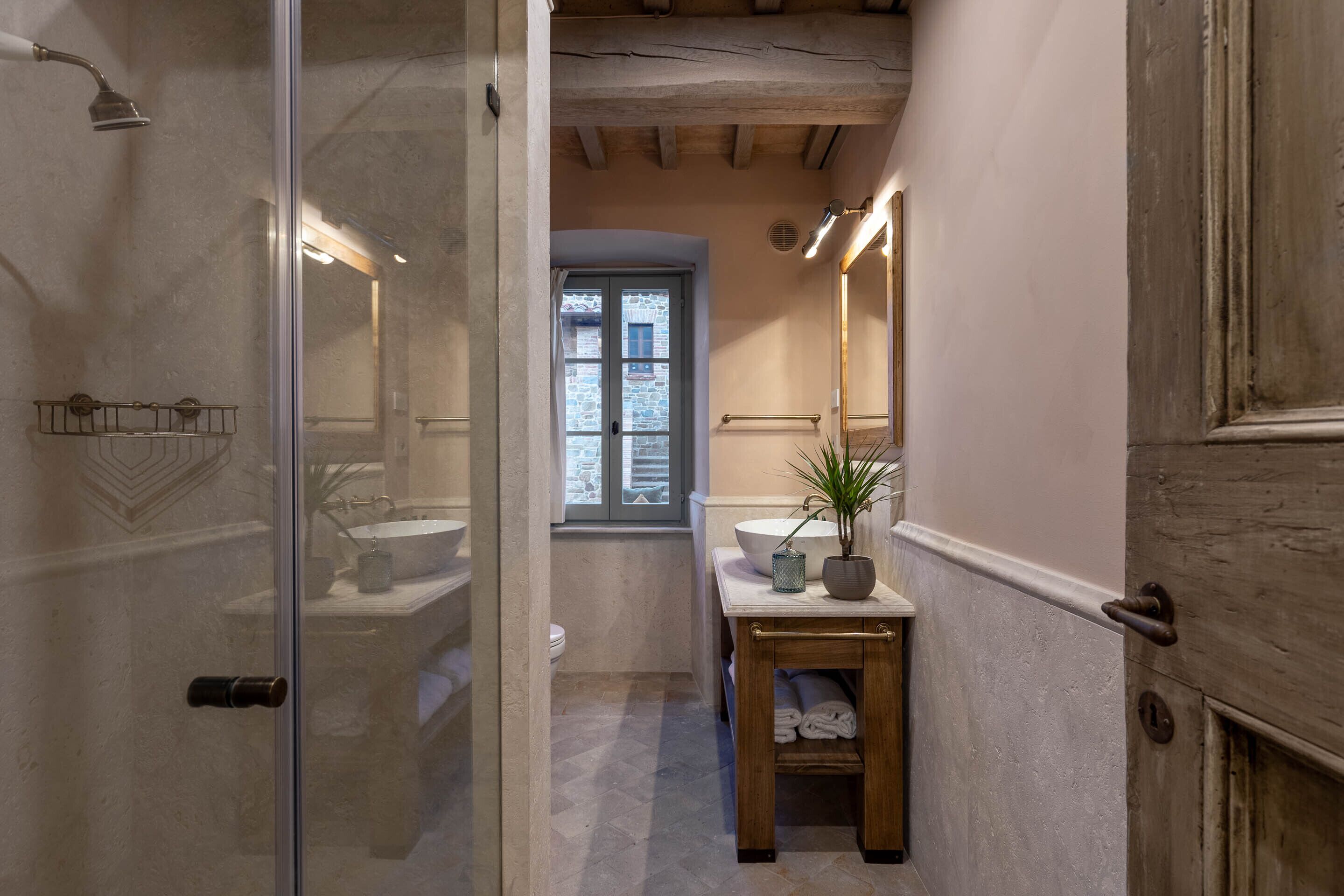 Un baño pequeño con puerta de vidrio, tocador de madera, lavamanos y ventana.