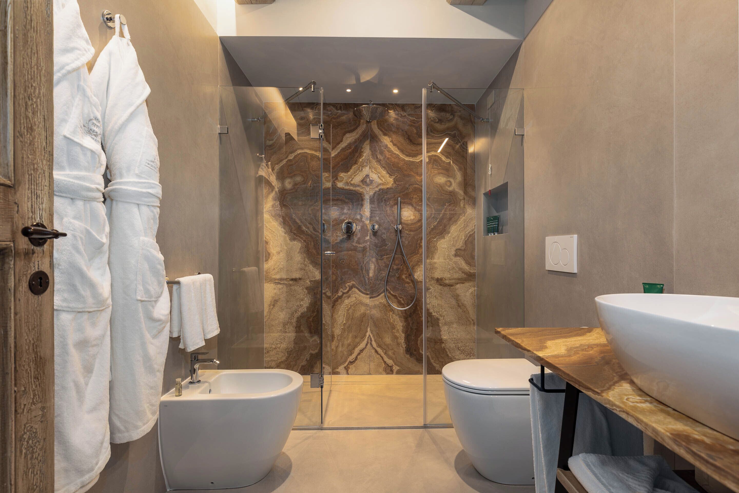 Ein modernes Badezimmer mit Marmorwand, einer Duschkabine aus Glas, zwei weißen Toiletten, einem hölzernen Waschtisch mit großem Becken und einem Handtuchhalter mit Handtüchern.