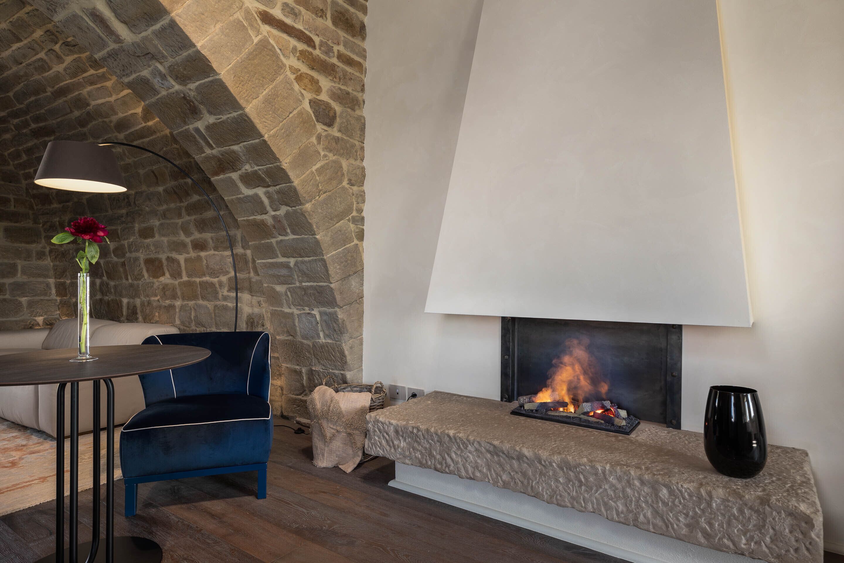 A cozy room with a stone fireplace, a blue chair, a round table, and a vase with a flower.
