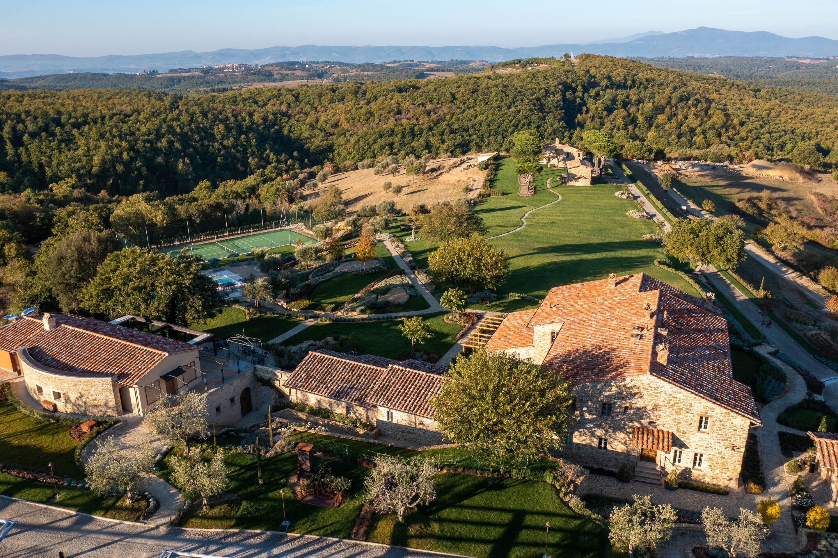 Vista aérea de una finca histórica con edificios de piedra, cancha de tenis y colinas boscosas circundantes.
