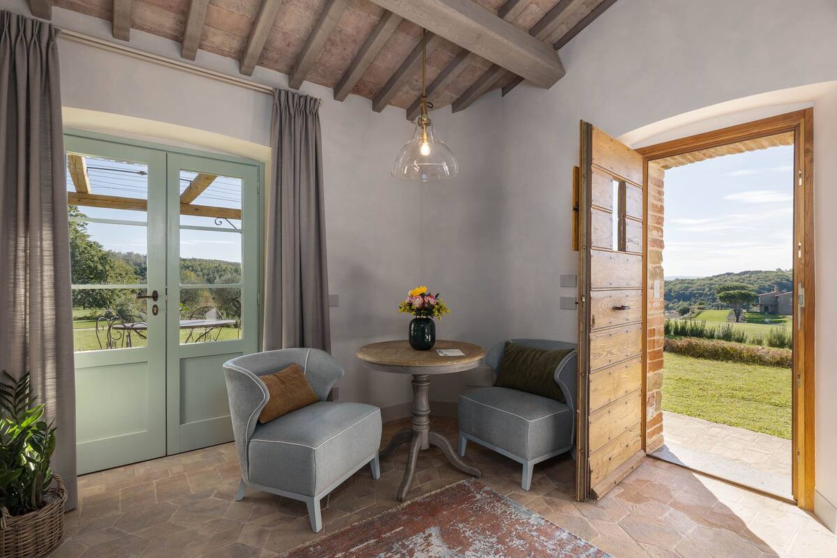 Una habitación con dos sillones grises, una mesa redonda de madera con un jarrón de flores, una lámpara colgante y vistas a un paisaje verde a través de una puerta.