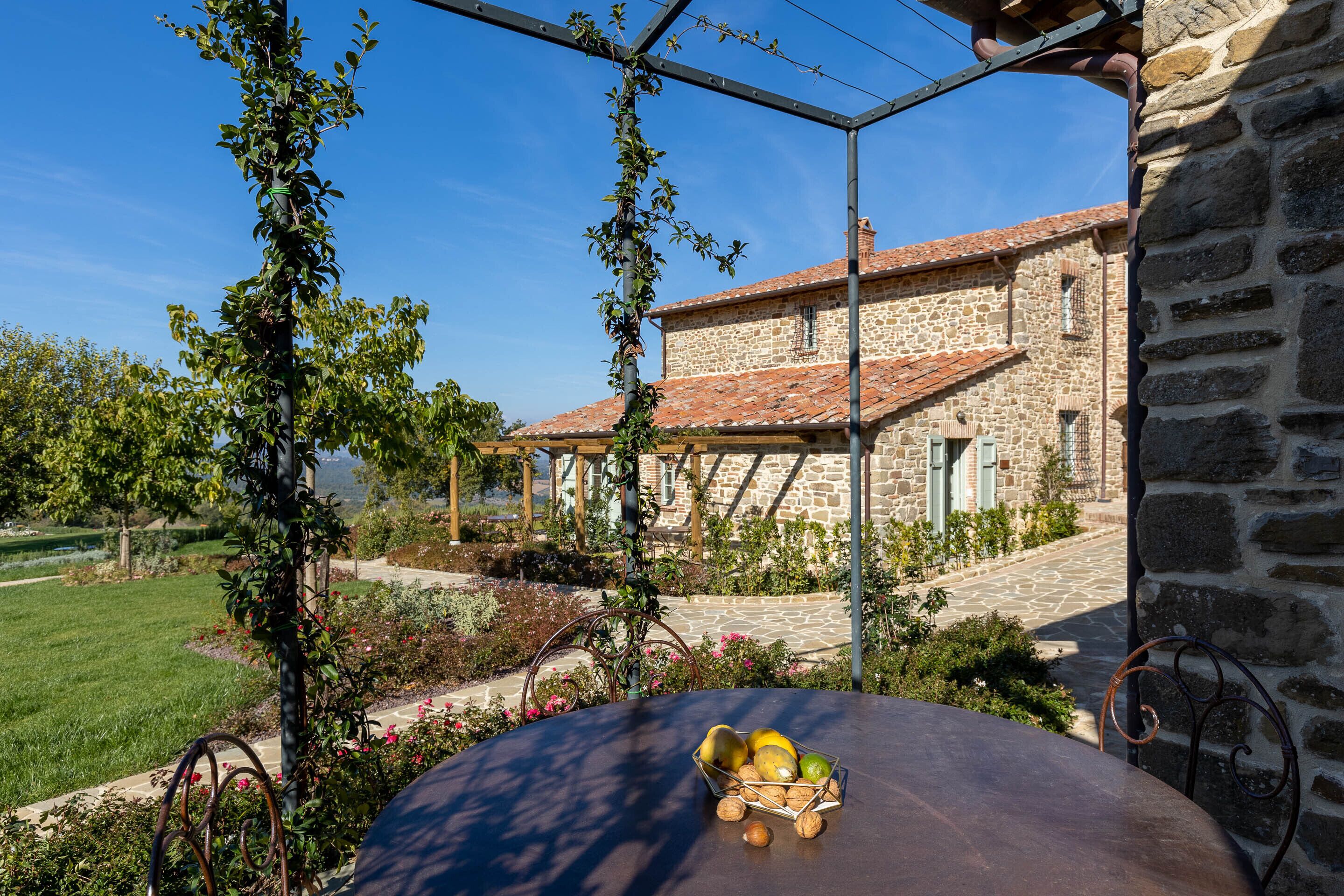 A stone building with a tiled roof, surrounded by a garden with a table featuring fruit and nuts.