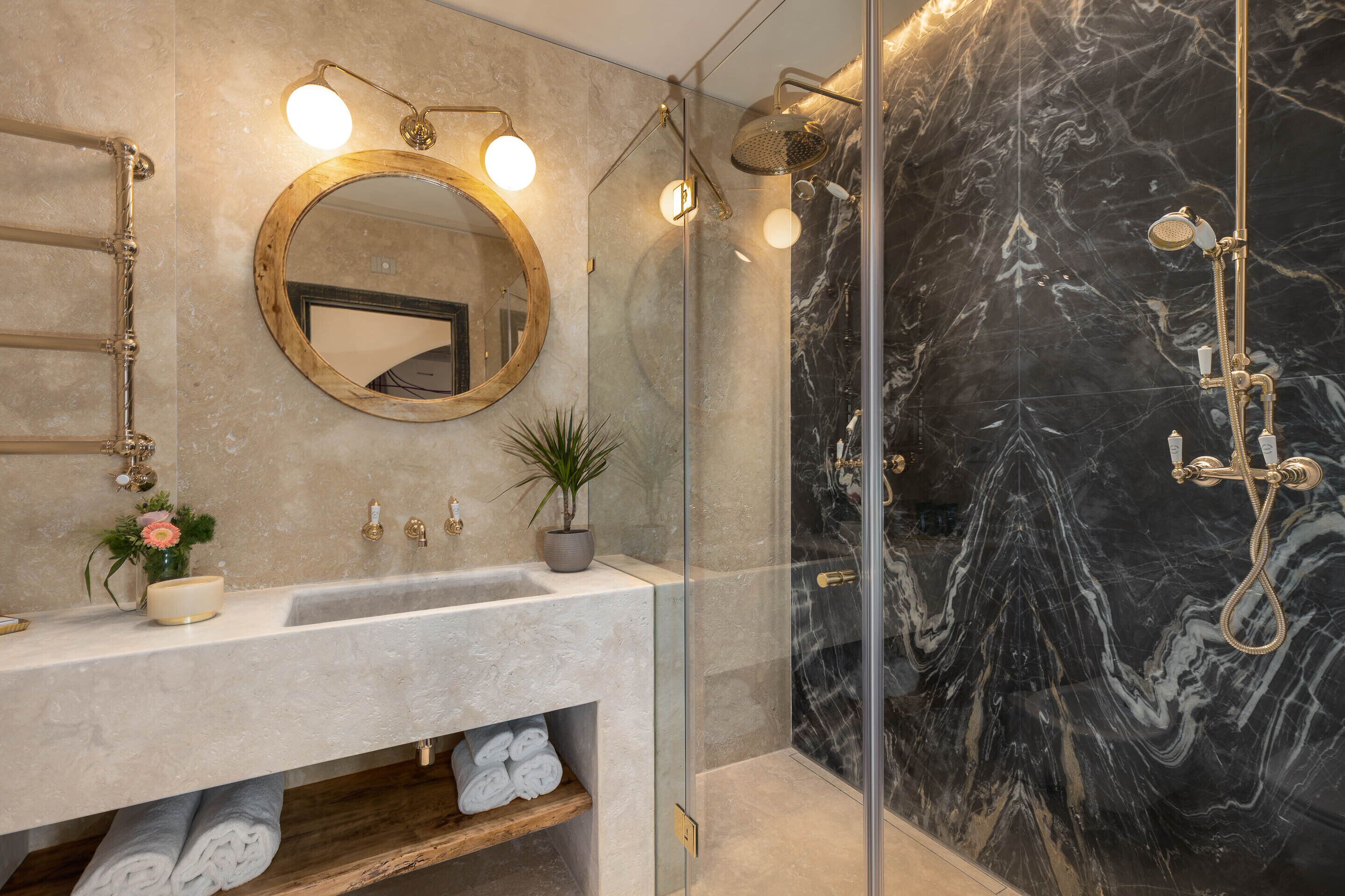 Un baño con pared de mármol, espejo redondo, lavamanos con planta y ducha con puerta de cristal.