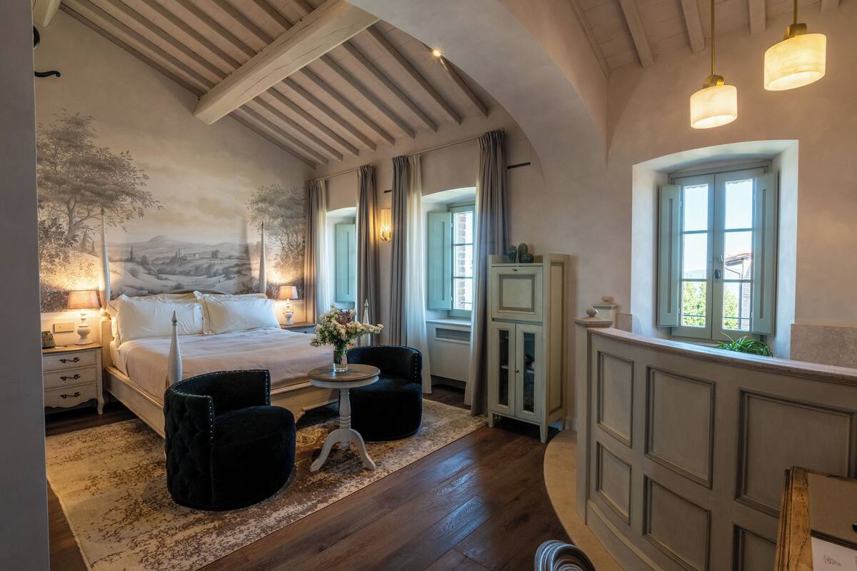 Un dormitorio con una cama grande, una mesita de noche, una silla, una pequeña mesa con un jarrón, y vistas al exterior a través de una ventana.