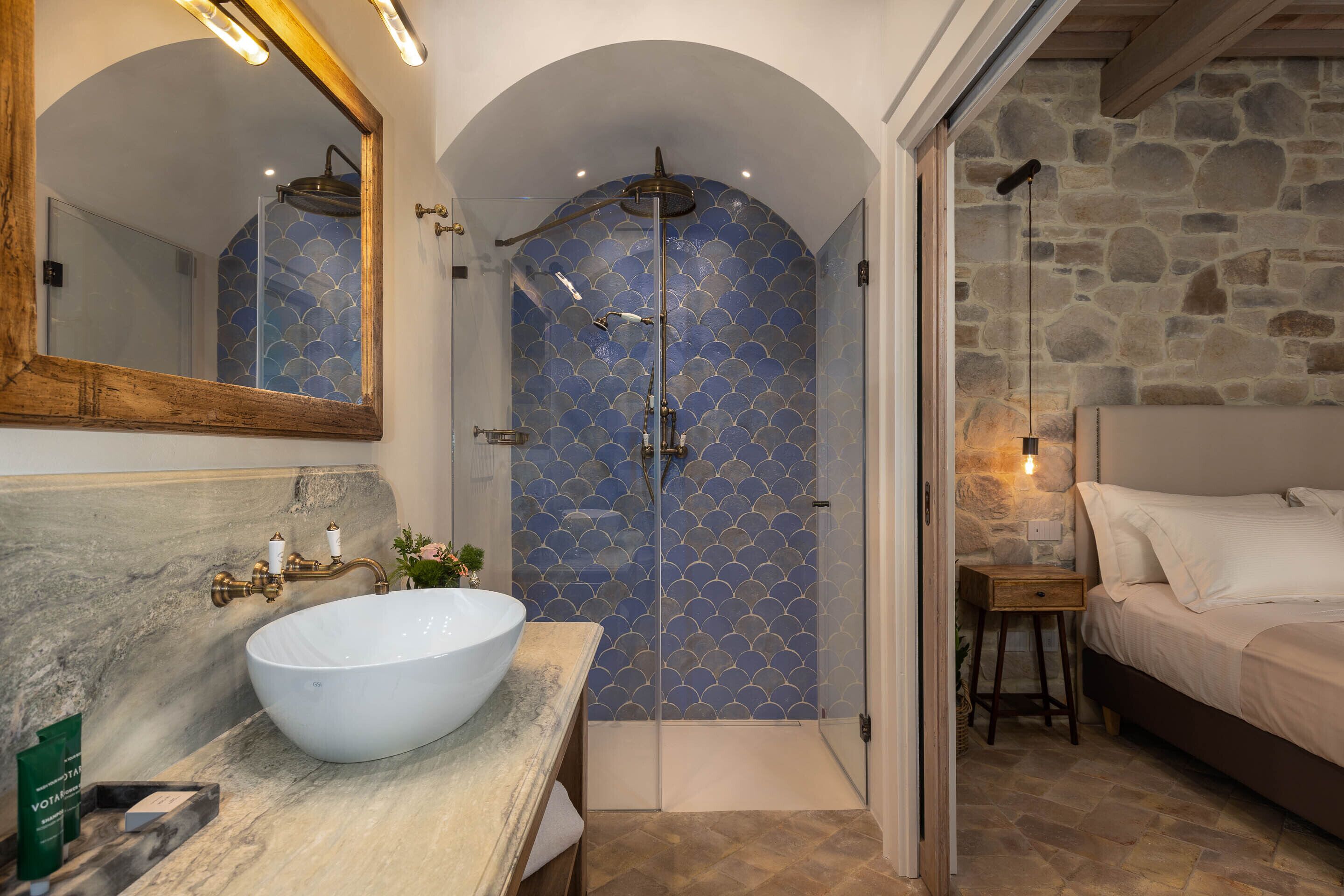 A bathroom with a large white vessel sink, a mirror, a shower with a blue tiled wall, and a bed with white linens.
