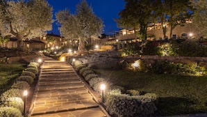 Un sendero de piedra que conduce a un edificio de piedra con múltiples ventanas y un jardín bien iluminado con setos y árboles cuidados.
