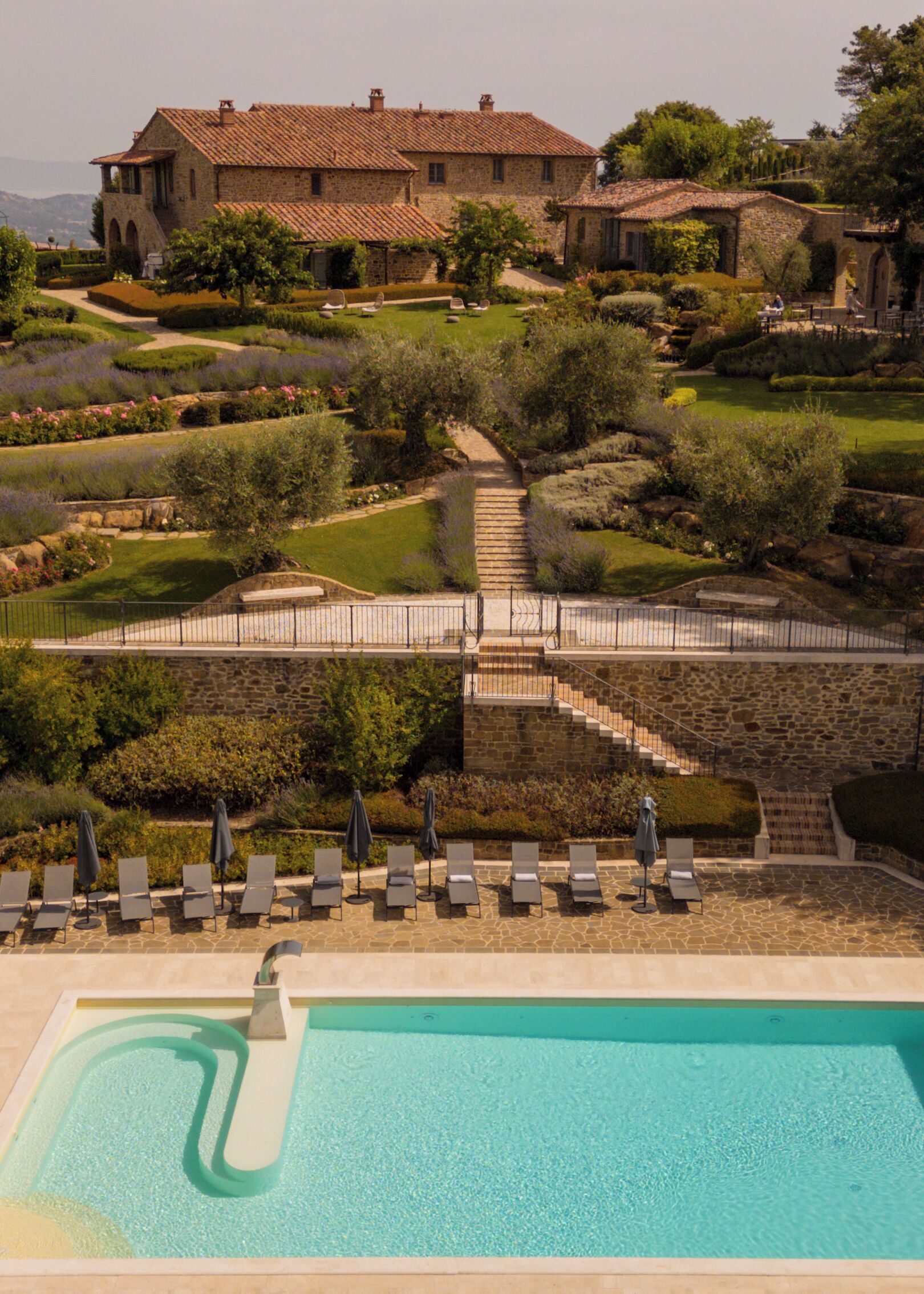 Zona de piscina con sillones de descanso y una escalera de piedra que conduce a un edificio con tejado de azulejos.