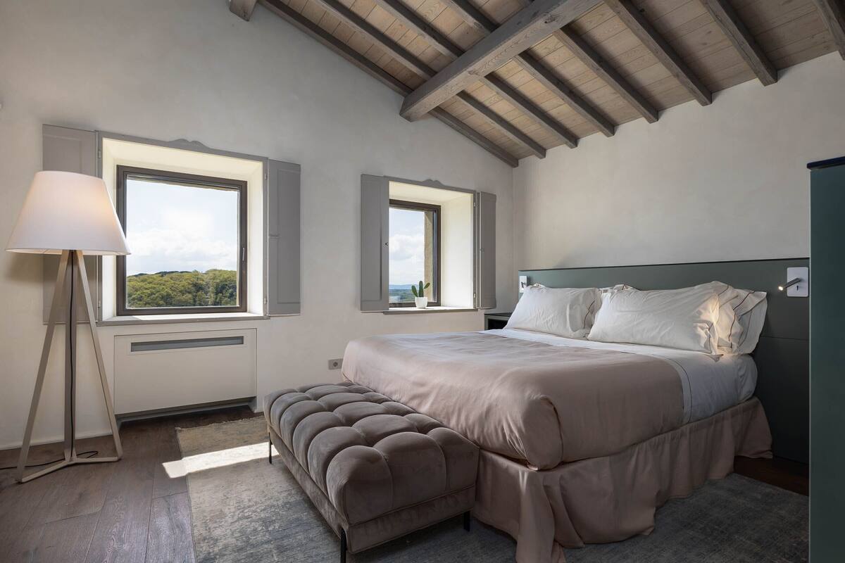 Un dormitorio con una cama, dos ventanas, una mesita de noche, una lámpara y un banco.