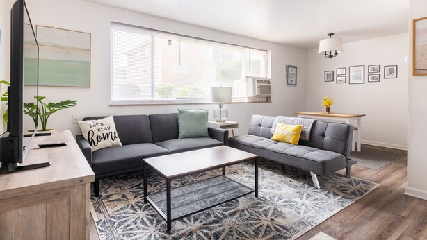 Een moderne woonkamer met een hoekbank, een salontafel en een televisie op een houten dressoir. Er is een raam met zonwering, een lamp en een bijzettafeltje met een bloemenvaas.