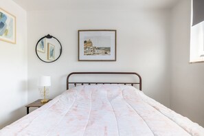Un lit bien fait, recouvert d’une couvre-lit à motifs roses et blancs, une table de chevet avec une lampe et un tableau encadré accroché au mur.