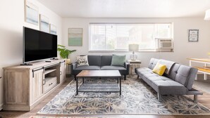 Een moderne woonkamer met een flatscreen televisie, grijze banken, een salontafel en een kamerkleed met een patroon.