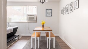 Een kleine, goed verlichte kamer met een houten tafel en bankjes, een raam met zonwering, een airconditioning en een muur met ingelijste foto’s.