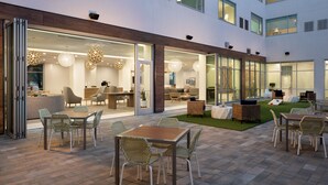 A modern outdoor patio area with seating arrangements, a pool, and a well-lit interior space visible through large windows.